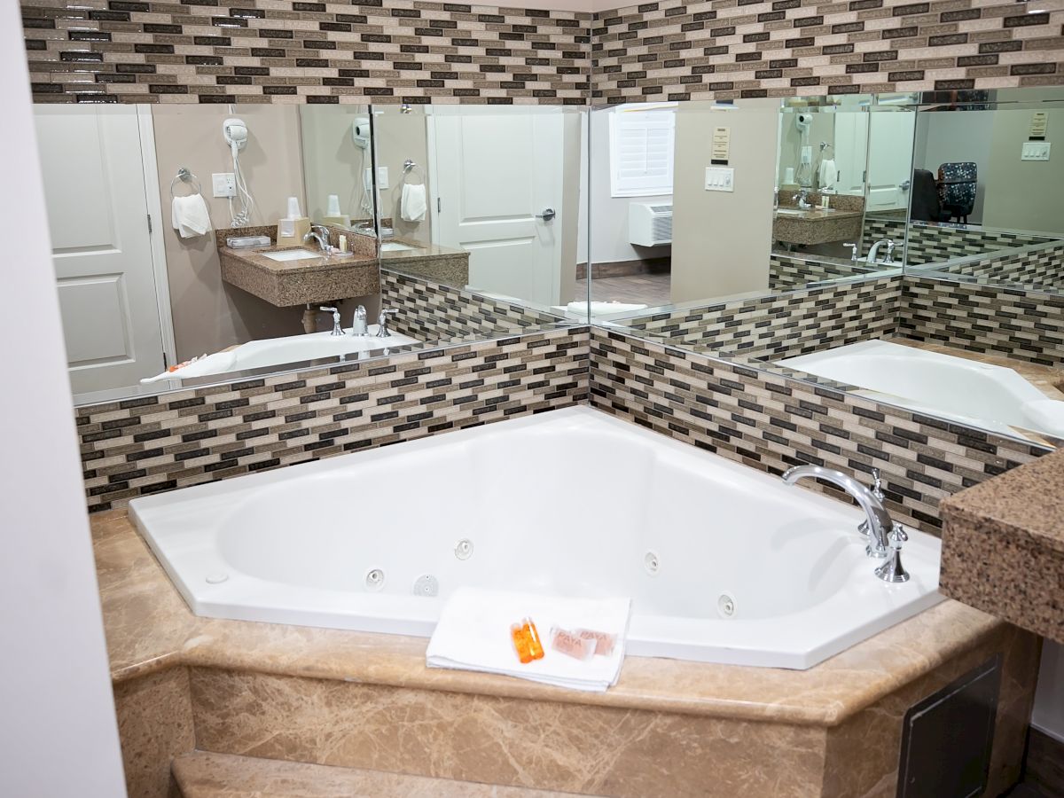 A modern bathroom featuring a corner jacuzzi tub, mosaic tile backsplash, mirrors, and a towel with an orange soap bar on the rim.