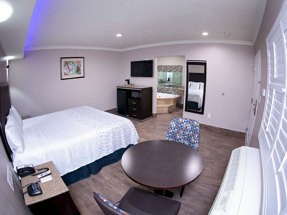 The image shows a modern hotel room with a double bed, a round table with chairs, a dresser with a TV, and an open bathroom door.