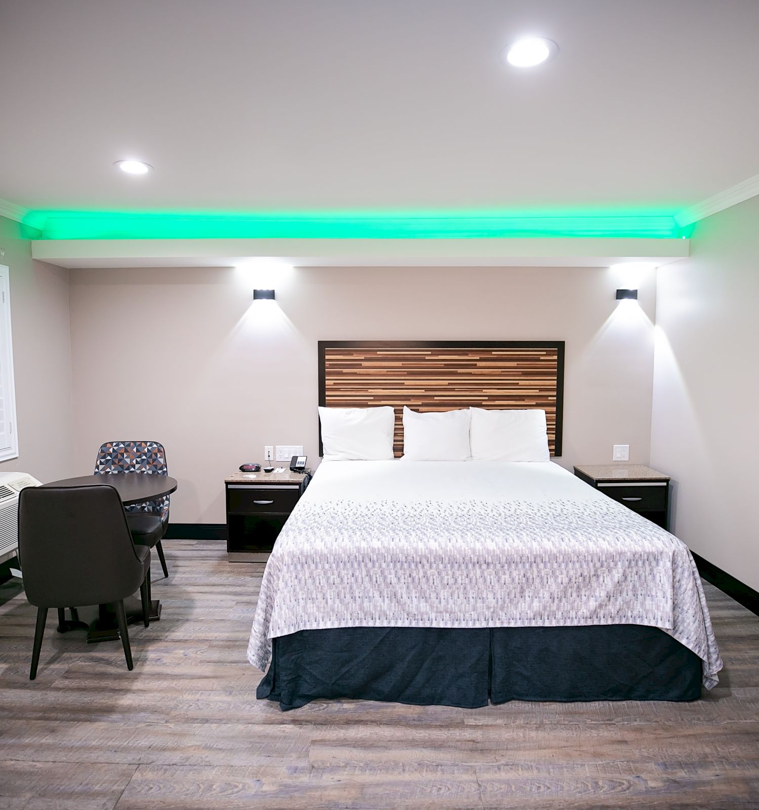 A modern hotel room with a bed, small desk with chair, wall art, and green accent lighting. Light-colored walls and wooden floors enhance the space.
