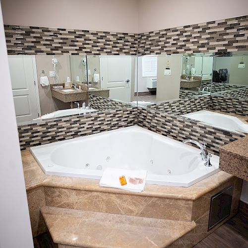 The image shows a luxurious bathroom featuring a corner whirlpool bathtub, marble tiles, a neatly arranged towel with toiletries, and large mirrors.