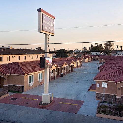 The image shows a row of single-story buildings with red roofs, resembling a modern motel or apartment complex, with a sign on a pole in the foreground.