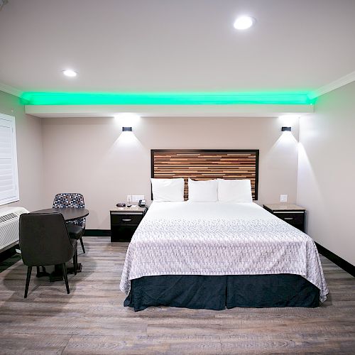 This image shows a modern hotel room with a bed, nightstands, a desk, chairs, and wall art, complemented by green LED accent lighting.