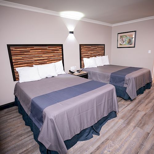This image shows a hotel room with two double beds, modern decor, wooden flooring, a wall-mounted AC unit, and a small artwork on the wall.