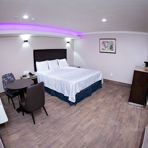 A modern hotel room features a large bed with white linens, a small table and chairs, artwork on the wall, and ambient purple lighting.