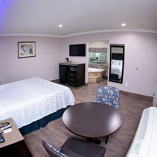 A modern hotel room features a bed, a round table with chairs, a TV, dresser, and a visible bathroom area with a bathtub in the background.