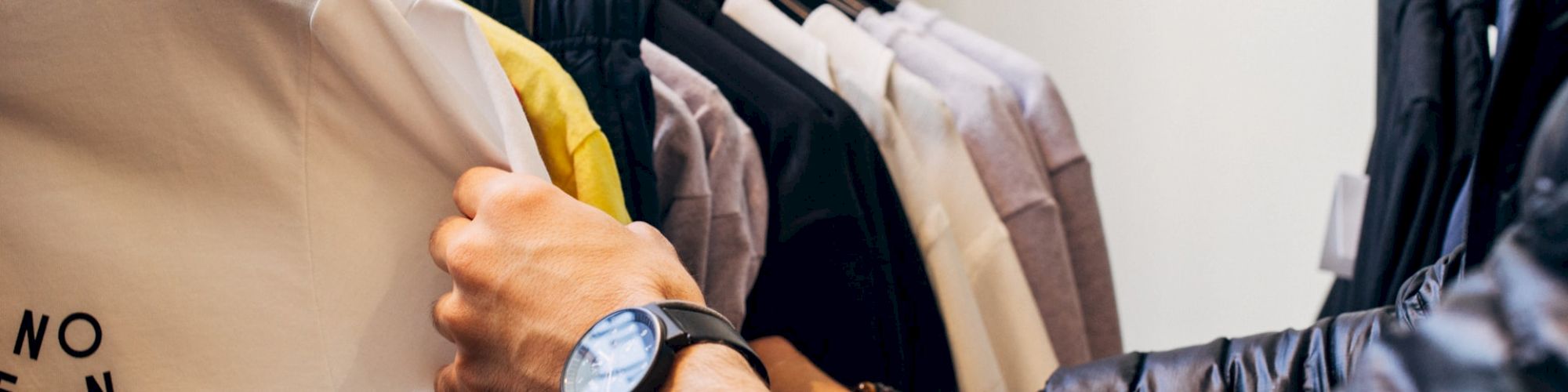 A person wearing a wristwatch is shopping for clothes, with their hands sifting through various colored shirts on a clothing rack.