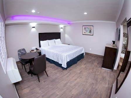 A modern hotel room with a large bed, two chairs, a small table, a dresser, and wall decor. The lighting includes a purple accent.